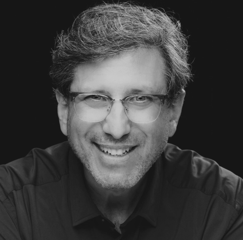 A black and white, close-up portrait of a smiling man with a light complexion. He has wavy, salt-and-pepper hair, short facial stubble, and is wearing clear-rimmed glasses. He is dressed in a dark button-down shirt, and the background is solid black.